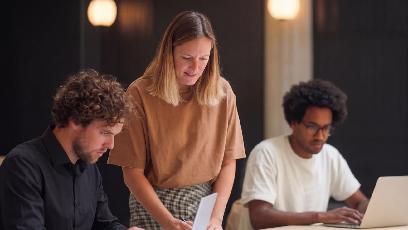 Person standing beside coworkers as they review information on laptops.