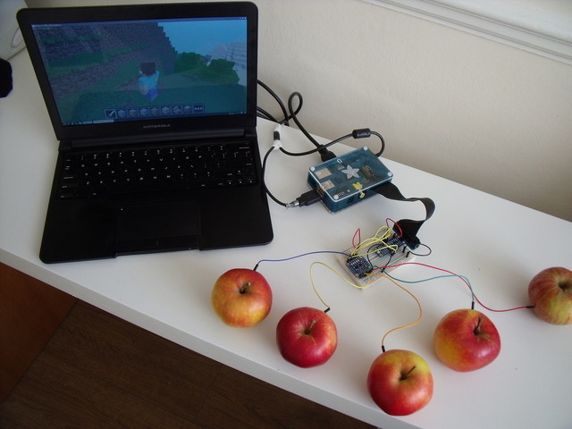 A laptop displays a game, connected to a Raspberry Pi in a clear case. The Pi connects to circuit board on a white breadboard with wires attached to apples, used as input devices. 