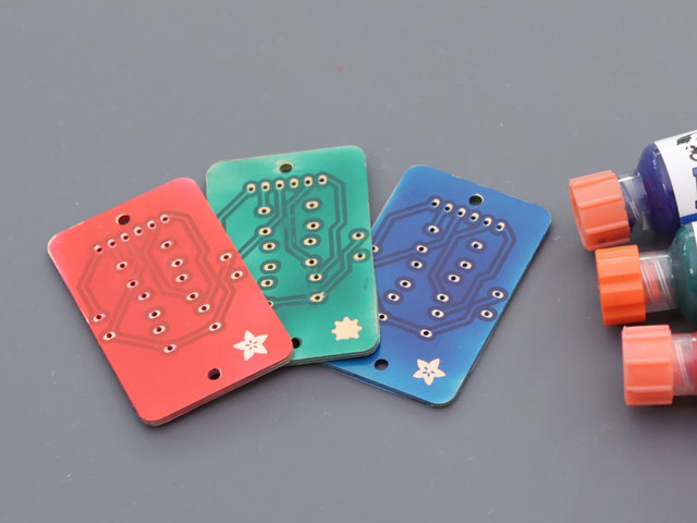 Three colorful rectangular circuit boards with visible solder points are arranged on a gray surface. Bottles of solder mask with orange caps are partially visible on the right.