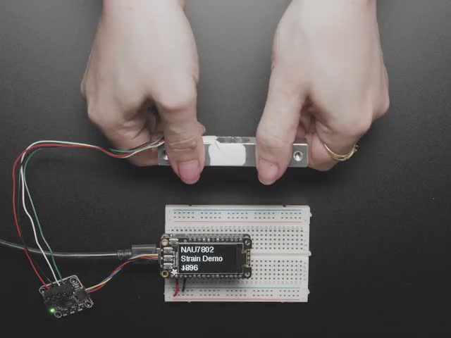 Adafruit NAU7802 24-Bit ADC - STEMMA QT / Qwiic - Video of a pair of white hand's trying to bend a strain gauge. The gauge is connected to a breakout board which is also connected to an OLED display on a half-size breadboard. The numbers on the OLED display show high digit numbers.