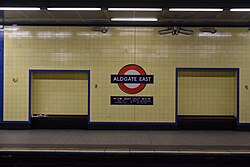 Platform rear wall with bright cream tiles and blue tiles used sparingly to mark details. A few relief tiles show icons and simple designs. There are two wooden bench seat recessed into alcoves and a large "Aldgate East" roundel with way-finding to the two station ends.