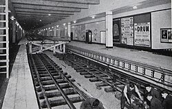 Black and white photo of station with workmen attending track. In the background the track is raised on wooden trestles at the same level as the new platforms and in the foreground it has been lowered to the correct level.